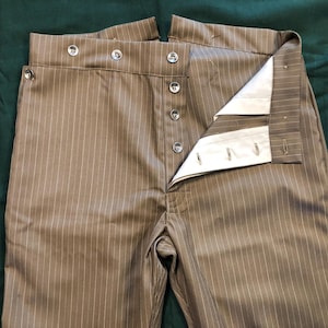 Puede incluir: Pantalones a rayas marrones y blancas con cintura alta y cierre de botones. Los pantalones están abiertos, revelando el forro interior y los detalles de los botones. La tela tiene un patrón de rayas verticales.