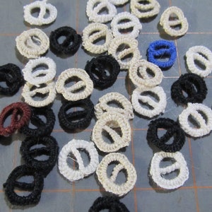 May include: A collection of small, circular, woven rope embellishments in various colors. The colors include off-white, black, burgundy, and blue. These decorative elements are likely used for crafts or sewing projects.
