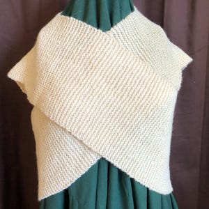 May include: Cream-colored knitted wrap with a textured, diagonal stitch pattern. The wrap is draped over a green garment, creating a layered effect. The fabric appears soft and warm, suitable for cooler weather.