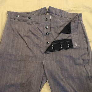 Puede incluir: Pantalones grises de raya diplomática con bragueta de botones y botones negros. Los pantalones están desabrochados, revelando la construcción interior. La tela tiene rayas verticales.