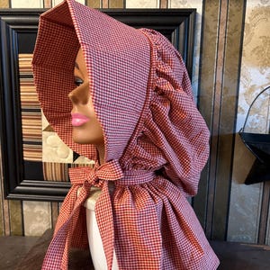 May include: A red and white gingham bonnet with a wide brim and ties. The bonnet is displayed on a mannequin head. The fabric has a classic checkered pattern, and the ties are knotted in a bow under the chin. The bonnet is a vintage style.