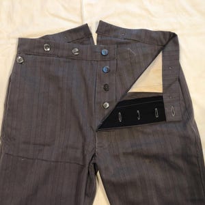 May include: Close-up of gray pinstripe trousers with button fly and side button details. The pants are unbuttoned, revealing the interior construction. The fabric has vertical stripes. The garment is likely vintage or historical.
