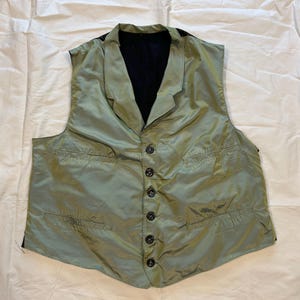 May include: A sleeveless vest in a light sage green color featuring a notched lapel collar, a dark interior lining, and a front button closure with six dark buttons. The garment includes two horizontal welt pockets on the front panels.