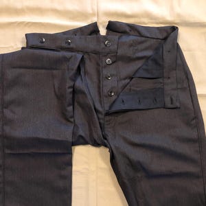 Puede incluir: Pantalones de raya diplomática gris oscuro con bragueta de botones y cierre de botón en la cintura. Los pantalones están colocados planos sobre una superficie beige, mostrando los detalles de la tela y la confección. Diseño clásico y a medida.