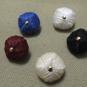 May include: Five colorful, round buttons in shades of blue, white, black, burgundy, and cream. Each button has a gold-colored center. The buttons are made of a woven material.