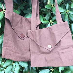 Pode incluir: Duas sacolas marrons com bolsos com aba e fechos de botão. As sacolas têm alças longas e são feitas de um tecido macio e texturizado. As sacolas são exibidas em um fundo de folhas verdes.