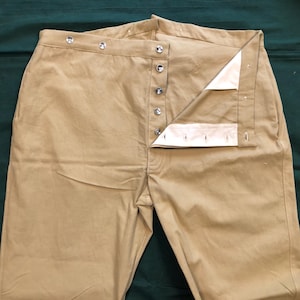Puede incluir: Pantalones de color beige con bragueta abotonada y varios botones a lo largo de la cintura. Los pantalones están hechos de una tela resistente y parecen ser de estilo vintage o histórico. La bragueta está parcialmente desabrochada, revelando un forro blanco.
