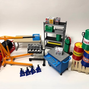 Puede incluir: Una escena de garaje en miniatura con varias herramientas y equipos. Incluye un compresor de aire rojo, un elevador de motor, un banco de trabajo, un carro de herramientas y varios barriles de aceite. También cuenta con una estantería con cajas de aceite y una caja verde.