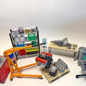 Puede incluir: Un conjunto de taller de garaje en miniatura con una grúa de motor amarilla levantando un bloque de motor negro y rojo. Incluye un carro de herramientas rojo, estantería con botellas de aceite, banco de trabajo con transmisión, piezas en palé y tanques de gas.