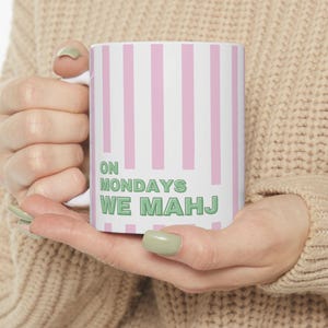 May include: White ceramic mug with pink vertical stripes and the text "ON MONDAYS WE MAHJ" in green. The mug is held by a person wearing a beige sweater. The mug is a standard size for coffee or tea.