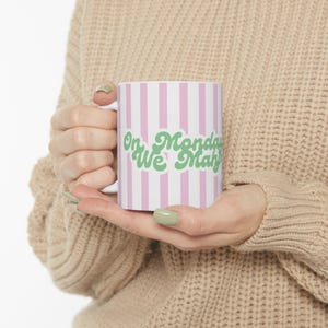 May include: White ceramic mug with pink vertical stripes and the text "On Monday We Mah". The text is in a green, retro-style font. The mug has a white handle and is held by a person.