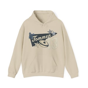 May include: A cream-colored hoodie with a front pocket and a drawstring hood. The front features a graphic with the word "Tommen", a shamrock, stars, and the number "13". The graphic has blue, white, and cream accents.