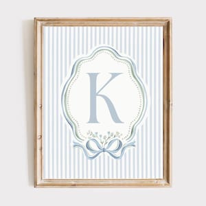 以下が含まれることがあります： 装飾的な楕円の中に、水色の文字「K」が描かれた額入りのアートワーク。背景は青と白のストライプ。下部には花のアクセントが付いた青いリボンがあります。フレームは明るい茶色です。