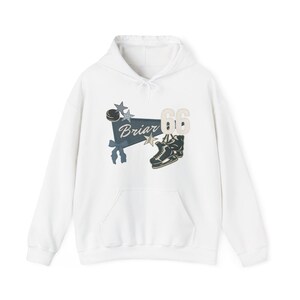 Puede incluir: Sudadera con capucha blanca con bolsillo delantero y capucha con cordón. El diseño presenta una pancarta azul con la palabra "Briar", un disco de hockey, estrellas, el número "66" y un par de patines de hielo.