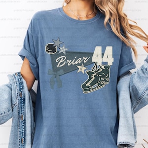 Op de afbeelding: Blauw T-shirt met een hockeythema. Het ontwerp bevat een hockeypuck, sterren, een banner met de naam "Briar", het nummer "44" en een hockeyschaats. Het shirt heeft opgerolde mouwen.