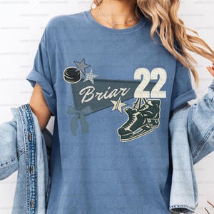Puede incluir: Camiseta azul claro con un diseño de hockey, que incluye un disco, estrellas y la palabra "Briar" con el número "22". La camiseta también presenta un gráfico de patines de hielo.