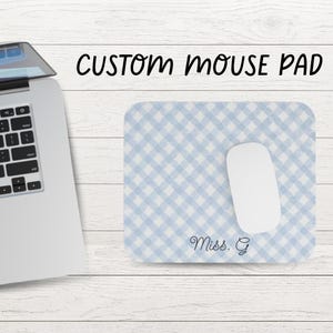 May include: A rectangular mouse pad featuring a light blue and white diagonal gingham pattern. The text "Miss. G" is printed in a dark blue cursive script in the bottom right corner. A white computer mouse rests on the surface of the mouse pad.