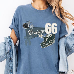 Puede incluir: Camiseta azul con un diseño temático de hockey. La camiseta presenta la palabra "Briar" en cursiva, el número "66", un patín de hockey, un disco y estrellas. La camiseta tiene las mangas enrolladas.