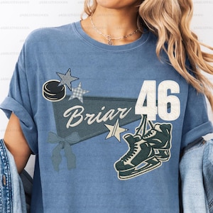May include: A light blue t-shirt featuring a hockey-themed design. The design includes a hockey puck, stars, the word "Briar", the number 46, and a pair of ice skates. The graphics are in shades of blue, white, and green.