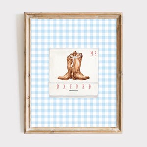 Puede incluir: Impresión en acuarela enmarcada con botas de vaquero marrones con un lazo blanco, sobre un fondo de cuadros azules y blancos. El texto "MS" y "OXFORD" está impreso en rojo. El marco es de color marrón claro.