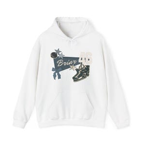 Puede incluir: Sudadera con capucha blanca con bolsillo delantero y cordón. El gráfico frontal presenta una pancarta con la palabra "Briar", el número "46", un disco de hockey, estrellas y un patín de figura.