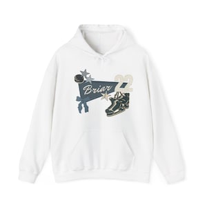 Puede incluir: Sudadera con capucha blanca con bolsillo delantero y diseño gráfico. El diseño incluye la palabra "Briar", un disco de hockey, estrellas, una pancarta, el número "22" y un par de patines de hielo.