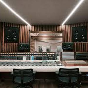 May include: A professional recording studio with a large mixing console, two black studio chairs, and two large speakers mounted on the wood-paneled walls. The room has a modern design with recessed lighting.