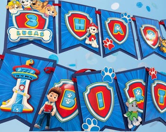 May include: A Paw Patrol themed birthday banner featuring blue pennants with red shield emblems, letters spelling out HAPPY BIRTHDAY, the name LUCAS, the number 3, and various character cutouts including Ryder, Marshall, and Rocky.