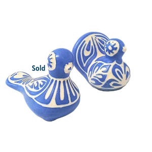 Puede incluir: Dos figuras de pájaros de cerámica azul con patrones decorativos blancos. Un pájaro está erguido y el otro está acostado. La palabra "Sold" es visible en la imagen.
