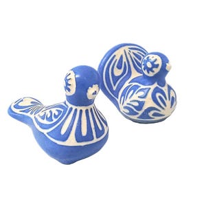 Puede incluir: Dos figuras de pájaros azules de cerámica con patrones decorativos blancos. Un pájaro está en posición vertical y el otro está sentado. Las figuras tienen un acabado brillante y están sobre un fondo blanco.