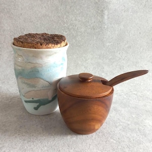 May include: A ceramic jar with a cork lid and a small wooden bowl with a lid and spoon. The jar has a white base with blue and tan watercolor-like designs. The wooden bowl is a warm brown color.
