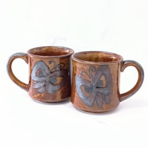 May include: Two brown ceramic mugs featuring a hand-painted grey butterfly design on the front. Each mug has a rounded handle and a smooth, glossy finish. The mugs are positioned side-by-side, showcasing the consistent earthy colour palette.