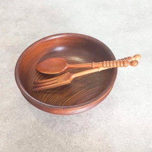 Könnte beinhalten: Eine Holzschüssel mit einem Löffel und einer Gabel. Die Schüssel ist rund und hat eine dunkelbraune Oberfläche. Löffel und Gabel sind hellbraun und haben dekorative Griffe. Das Set ist zum Servieren oder Essen gedacht.