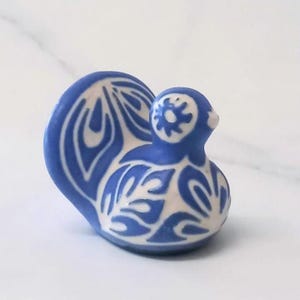 Puede incluir: Una pequeña figura de cerámica con forma de ave, cuerpo redondeado y cola en abanico. La pieza presenta un diseño bicolor con base blanca y complejos patrones florales y de hojas en color azul que cubren toda la superficie del objeto.