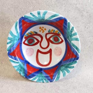 May include: A white ceramic plate with a hand-painted sun face design. The sun features red, blue, and teal accents, with a smiling mouth and rosy cheeks. The plate's rim has a teal and blue pattern.