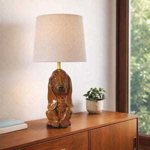 May include: A table lamp featuring a glossy brown ceramic base shaped like a seated basset hound dog. The lamp has a gold-toned metal neck and a simple, tapered, light beige fabric drum lampshade that sits atop the dog-shaped ceramic base.