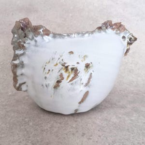 Puede incluir: Un cuenco de cerámica blanca hecho a mano con una forma orgánica única. El cuenco presenta un borde texturizado y rugoso con detalles marrones y sutiles marcas marrones en la superficie. El interior es liso y esmaltado.