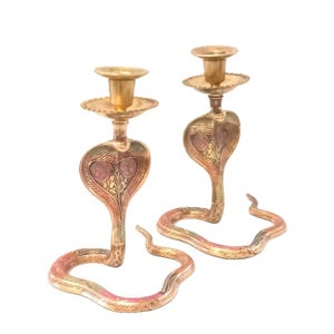 Könnte beinhalten: Zwei messingfarbene Kerzenhalter in Kobraform. Die Kerzenhalter haben eine goldfarbene Oberfläche mit roten und rosa Akzenten. Die Kobraköpfe sind mit geätzten Mustern versehen. Die Kerzenhalter sind für eine einzelne Kerze konzipiert.