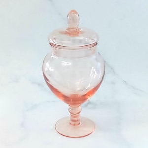 May include: A translucent pale pink glass jar with a rounded body, a matching removable lid featuring a small knob handle, and a pedestal base. The glass has a smooth, polished finish and a consistent light pink hue throughout the entire structure.