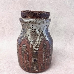 May include: A handmade ceramic jar with a cork stopper. The jar has a speckled white and brown glaze with reddish-brown abstract designs. The dark brown cork stopper fits securely on the jar's opening. The jar is a decorative storage container.