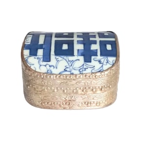 Puede incluir: Pequeña caja rectangular para baratijas con base de metal plateado y tapa de porcelana. La tapa presenta un diseño floral azul y blanco con caracteres chinos. La caja tiene un patrón decorativo y texturizado alrededor de los lados.