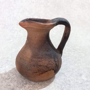 May include: A small, rustic terracotta jug with a handle. The jug has a rounded body and a flared spout. The colour is a mix of brown and black, with a textured surface. The handle is arched and attached to the top of the jug.