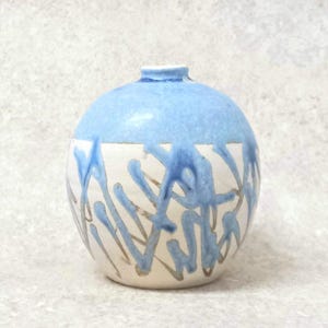 Puede incluir: Un pequeño jarrón de cerámica redondo con la parte superior azul claro y la base blanca. La base blanca presenta patrones de esmalte goteado azul y dorado. El jarrón tiene una abertura estrecha en la parte superior.