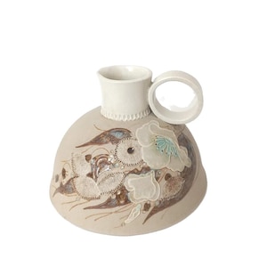 May include: A beige ceramic vase with a white rim and handle. The vase is decorated with floral and leaf designs in shades of brown, white, and teal. The vase has a unique shape and a textured surface.