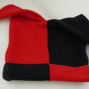 May include: A red and black jester hat with a square base and pointed tips. The hat is divided into four sections, with alternating red and black panels. Each tip has a small white bell with a smiling face.