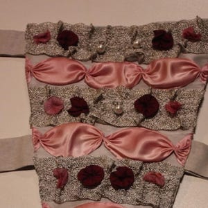 May include: A vintage-style corset in a muted color palette. The corset features horizontal bands of pink satin and decorative lace, adorned with small, dark red fabric flowers and faux pearls. Beige fabric extends from the sides.