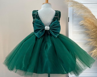 Op de afbeelding: Groene jurk met een grote strik op de rug, tentoongesteld op een houten mannequin. De jurk heeft een tule rok en een satijnen lijfje. De strik is versierd met een sprankelende versiering. De jurk is ontworpen voor een speciale gelegenheid.
