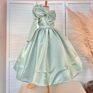 May include: A mint green satin dress with a large bow on the shoulder and a full skirt. The bow is adorned with small, pearl-like embellishments. The dress has one long sleeve and is displayed on a mannequin.