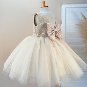 May include: A cream-colored formal dress with a tulle skirt and a large satin bow on the back. The bodice is embellished with delicate beadwork. The dress is displayed on a mannequin, showcasing its elegant design, perfect for special occasions.