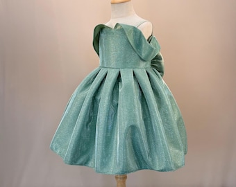 Op de afbeelding: Een mintgroene jurk met een glinsterende afwerking, tentoongesteld op een houten mannequin. De jurk heeft een geplooide rok en een unieke off-the-shoulder halslijn met een strik op de rug. De jurk is ontworpen voor een kind.